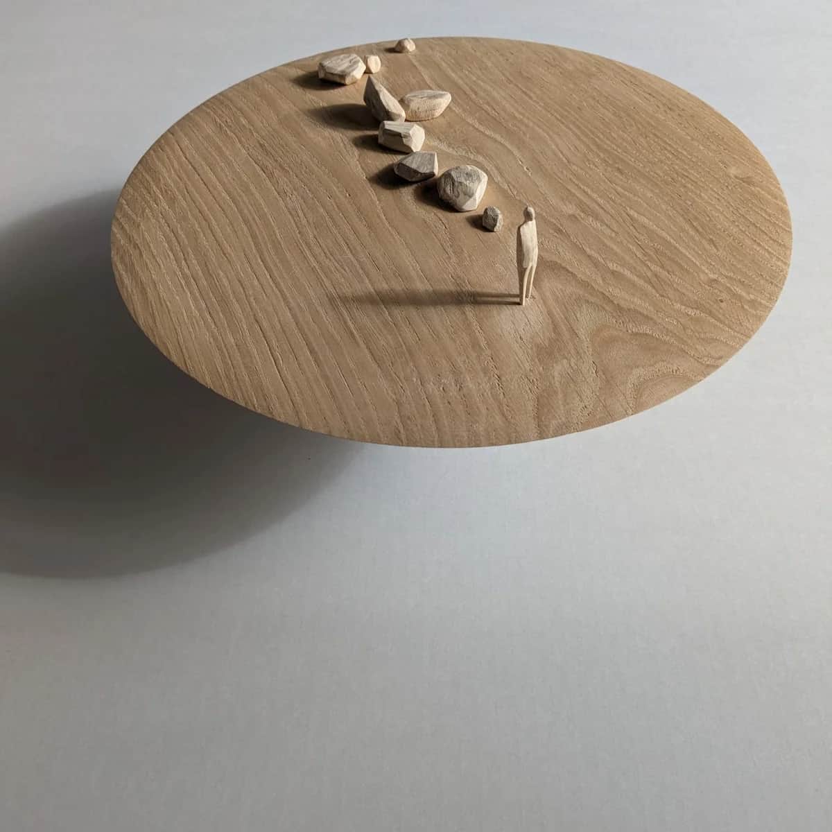 A carved oak disc holding small wooden rocks and a single standing figure, a sculpture by Ruth Howes